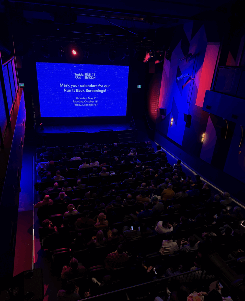 A dimly lit theater filled with people seated and facing a large screen displaying event details for upcoming "Run It Back Screenings". The walls are decorated with blue and purple lighting.